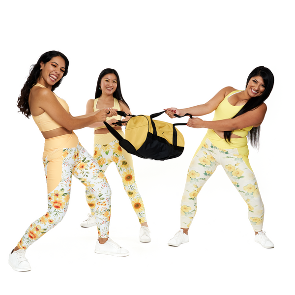 Three women in matching yellow and white outfits with sunflower patterns holding a black bag against a white background.